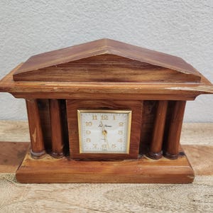 May include: A wooden mantel clock shaped like a temple with a triangular pediment and four columns. The clock face is square with a white background, gold-colored numbers, and gold hands. The text Seth Thomas is printed on the face.
