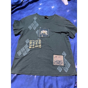 May include: A dark green short-sleeve t-shirt featuring light blue diamond-shaped patterns and three rectangular fabric patches. The patches display a white flower, gold Asian characters, and a beige graphic with waves and small red symbols.