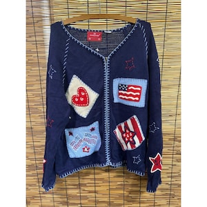 May include: A navy blue zip-up knit cardigan featuring a V-neckline and light blue whip-stitched edges. The garment is decorated with various embroidered patches, including a red heart, an American flag, and stars in red and white.