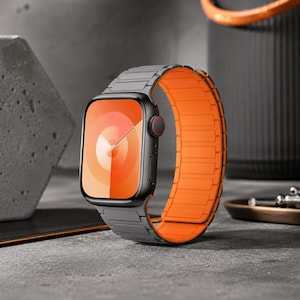 May include: A smartwatch with a rectangular black case and a digital crown featuring a red accent. The watch band is dual-toned, with a dark gray exterior and a vibrant orange interior, composed of segmented links that wrap around the device.