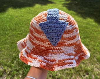 May include: A crochet bucket hat featuring a variegated orange and white yarn pattern. A prominent light blue arrow is stitched onto the center of the crown, extending from the top down toward the brim of the hat, crafted with visible yarn stitches.