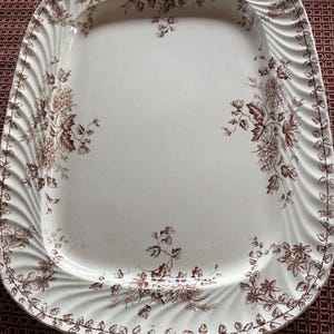 May include: A rectangular ceramic serving platter with rounded corners, featuring a cream-colored base and a brown floral transferware pattern. The rim has a raised, diagonal ribbed texture and a continuous vine border with floral clusters.