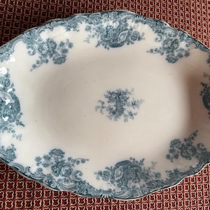 May include: An oval ceramic serving platter with a white base and a scalloped edge. The platter features a teal blue transferware pattern consisting of floral bouquets, scrolling foliage, and a central urn motif printed on the surface.