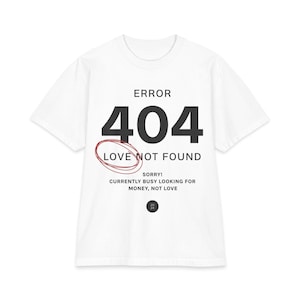 May include: A white short-sleeved t-shirt featuring black text that reads ERROR 404 LOVE NOT FOUND with a red hand-drawn circle around the word LOVE. Below this, the text reads SORRY! CURRENTLY BUSY LOOKING FOR MONEY, NOT LOVE and a small black circle with 20 24.