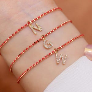 Puede incluir: Tres pulseras delicadas con bandas de cordón rojo fino y dijes de metal dorado con forma de las letras N, G y W. Cada dije de letra está incrustado con pequeñas piedras claras y brillantes que captan la luz, creando un aspecto superpuesto.