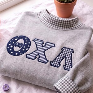 May include: A light gray sweatshirt featuring a mock neck collar with a navy and white gingham pattern. The front displays the Greek letters Theta Chi Alpha in navy blue with white star, stripe, and gingham patterns. The cuffs also feature gingham.