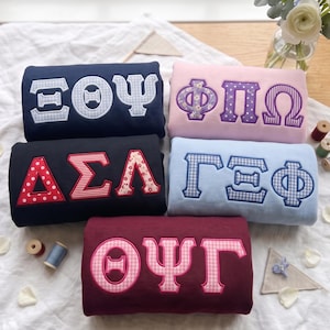 May include: Five folded sweatshirts in navy, light pink, black, light blue, and maroon. Each features large Greek letters appliqued in various patterns including gingham, polka dots, stripes, and floral prints, showcasing symbols like Xi Theta Psi, Phi Pi Omega, Delta Sigma Lambda, Gamma Xi Phi, and Theta Psi Gamma.