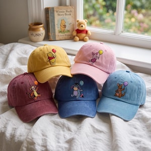 May include: Five baseball caps in yellow, pink, maroon, dark blue, and light blue. Each cap features an embroidered character holding a balloon: Winnie the Pooh with a red balloon, Piglet with a yellow balloon, Tigger with an orange balloon, Eeyore with a blue balloon, and Roo with a purple balloon.