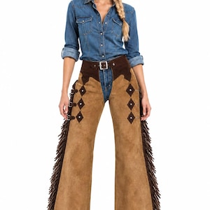 May include: Tan suede chaps featuring dark brown leather diamond-shaped accents, silver-toned buckles, and long fringe along the outer legs. The chaps are worn over a blue denim button-down shirt and blue jeans, paired with a brown cowboy hat.