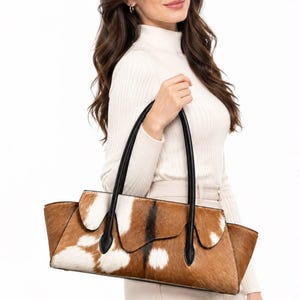 May include: A horizontal handbag featuring a brown and white cowhide pattern with a textured fur finish. The bag includes a structured flap closure, black leather trim, and two long, slender black leather shoulder straps for carrying.