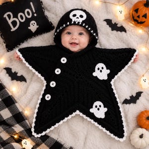 May include: A black star-shaped crochet baby bunting with a hood featuring a white skull motif. The garment has a white scalloped edge, four white buttons along the diagonal front, and two white ghost appliqués on the front surface.