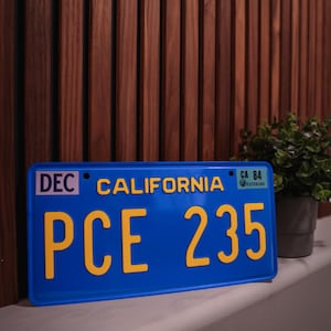 Può includere: Una targa rettangolare blu della California con caratteri gialli in rilievo che leggono PCE 235. La targa presenta il testo CALIFORNIA in giallo, un adesivo rosa DEC sulla sinistra e un adesivo verde CA 84 sul lato destro.