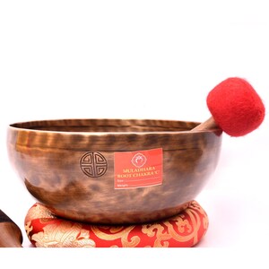 May include: A bronze-colored metal singing bowl with a hammered texture, featuring a circular emblem and a red sticker labeled MULADHARA ROOT CHAKRA C. A red felt-covered mallet rests inside the bowl, which sits on a red and gold patterned cushion.