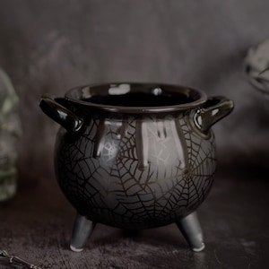 May include: A black ceramic cauldron with three short legs and two side handles. The exterior features a dark grey spider web pattern and a glossy drip design around the rim. The interior is smooth and black, with a wide circular opening.
