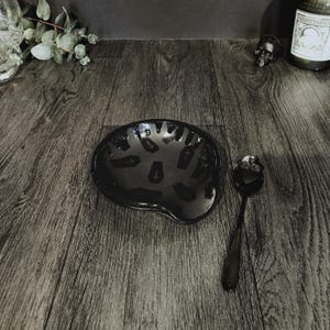 May include: A black ceramic spoon rest featuring a repeating pattern of small, dark grey coffin icons with cross details, accompanied by a matching black metal spoon with a skull-shaped handle, both designed for kitchen or dining utility.