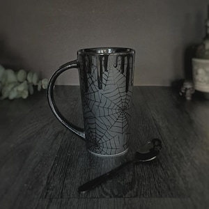 May include: A tall, cylindrical ceramic mug featuring a matte gray finish with a black spiderweb pattern and a black drip design around the rim. A matching black spoon with a skull-shaped handle rests on the surface next to the mug.