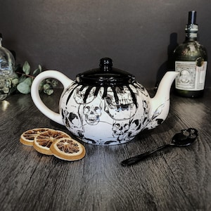 May include: A white ceramic teapot featuring a black skull pattern and a black drip design on the lid and upper body. The teapot has a curved handle, a spout, and a matching lid. Beside it are three dried orange slices and a black spoon.