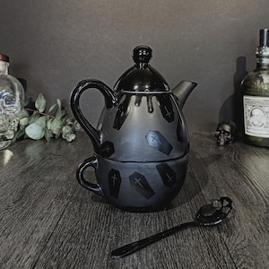 May include: A matte black ceramic tea set for one, featuring a teapot stacked on a teacup. Both pieces are decorated with a repeating pattern of glossy black coffin silhouettes. The set includes a matching black spoon with a skull-shaped handle.