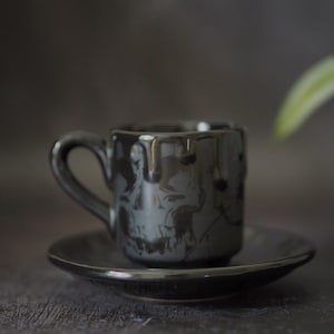 May include: A glossy black ceramic espresso cup and matching saucer. The cup features a dark grey skull graphic on the front and a decorative drip pattern around the upper rim. The handle is curved and integrated into the side of the cup.