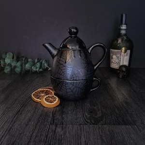 May include: A black ceramic tea-for-one set featuring a teapot that stacks onto a matching teacup. The surface of both pieces is decorated with a dark grey spiderweb pattern. The teapot has a lid with a round knob and a curved handle.