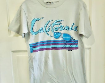 May include: A pale grey t-shirt with the word "California" in blue, designed to look like water, with blue sunglasses and a striped pattern. The t-shirt is hanging on a clear hanger.