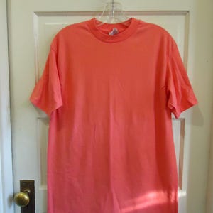 May include: A solid coral-colored t-shirt hangs on a clear plastic hanger. The short-sleeved shirt is made of a soft material and is displayed against a white door. The shirt has a crew neck and a simple, classic design.