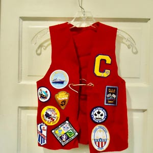 May include: Red vest with various embroidered patches, including a large "C" on the front, a "Junior Ranger Yellowstone" patch, and a "Pinewood Derby" patch.