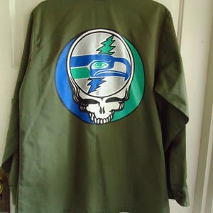 May include: Olive green button-up shirt with a large graphic on the back. The graphic combines a skull with a football team logo in silver, blue, and green. The shirt has long sleeves and a collar.