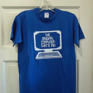 May include: Royal blue t-shirt with a white pixelated computer graphic and the text "THE ORIGINAL COMPUTER - THAT'S ME!". The shirt is hanging on a clear plastic hanger.