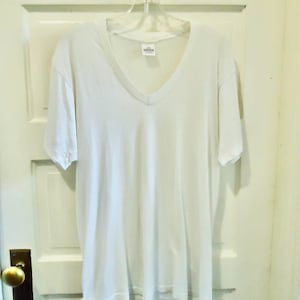 May include: A plain white V-neck t-shirt hanging on a clear plastic hanger. The shirt is made of a soft material and has short sleeves. The shirt is hanging on a white door.