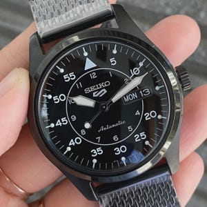 May include: A black Seiko 5 automatic wristwatch with a black dial featuring white Arabic numerals for minutes and hours, a day-date window at the three o'clock position, luminous hands, a triangular marker at twelve, and a metallic mesh strap.
