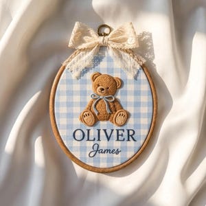 Puede incluir: Un bastidor de bordado ovalado de madera con un fondo de tela de cuadros vichy azul claro y blanco. Un oso de peluche marrón bordado con una cinta azul claro se sitúa sobre el texto OLIVER James. Un lazo de encaje crema está fijado arriba.