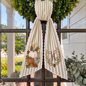 Puede incluir: Un colgador de puerta de tela decorativo con un patrón de rayas verticales en verde claro y blanco y bordes festoneados. Un lado muestra calabazas y hojas de otoño bordadas, mientras que el otro lado presenta una letra H monograma en una corona.