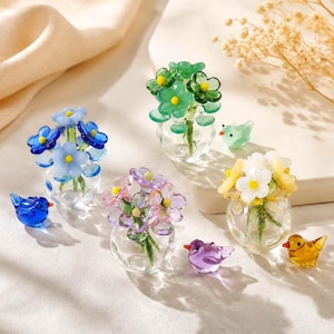 May include: Four miniature glass vases containing small bouquets of blue, green, pink, and white flowers, each accompanied by a tiny matching glass bird figurine in blue, green, purple, and yellow, arranged on a light surface.