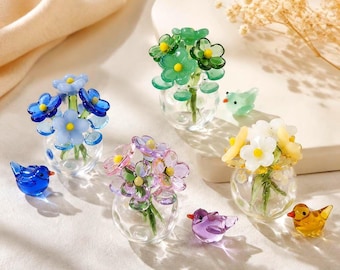 May include: Four miniature glass vases containing small bouquets of blue, green, pink, and white flowers, each accompanied by a tiny matching glass bird figurine in blue, green, purple, and yellow, arranged on a light surface.
