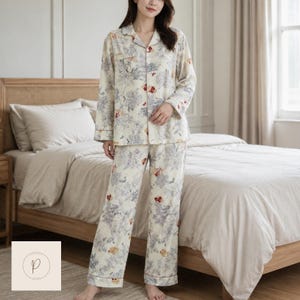 May include: A two-piece pyjama set consisting of a long-sleeved button-down shirt and matching trousers. The fabric is cream-coloured with a detailed toile-style print in shades of blue, grey, and red, featuring floral and botanical patterns throughout.