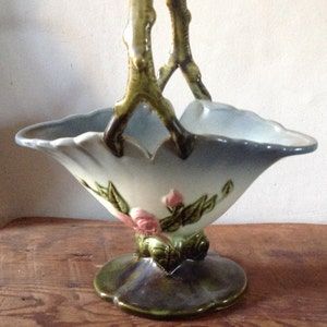 Puede incluir: Una cesta de cerámica vintage con un asa de rama verde y un diseño floral. La cesta es blanca con flores y hojas rosas y verdes. Tiene un esmalte azul en el interior y un esmalte verde en la base.