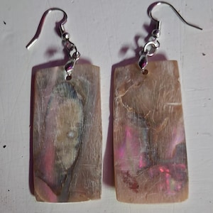 Può includere: Un paio di orecchini rettangolari con finitura perlescente e iridescente. Gli orecchini hanno un gancio e una catena color argento. La forma rettangolare ha un aspetto naturale e organico con sfumature rosa e viola.