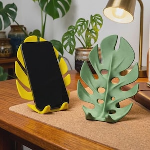 May include: A bright yellow monstera leaf-shaped phone stand holding a black smartphone, positioned next to a sage green monstera leaf-shaped phone stand. Both stands feature a cutout design mimicking the natural fenestrations of a tropical leaf.