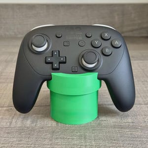 May include: A black Nintendo Switch Pro controller with white thumbstick accents resting on a bright green cylindrical pipe-shaped stand. The controller features standard A, B, X, and Y buttons, a directional pad, and two analog sticks.