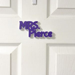 May include: A three-dimensional wall sign featuring the purple text "MRS. Pierce" in a bold, rounded font. The letters have a white border and are mounted on a white paneled door. The sign is positioned horizontally in the center of the frame.