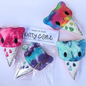 May include: Three handmade catnip toys shaped like ice cream cones. Each cone is a different color: pink, blue, and rainbow. The cones are made of fleece and have a cat face on the top. The text on the packaging reads "Organic & Vegan Kitty Cone Handmade of fleece, organic catnip & a touch of fiber fill. USDA Organic."