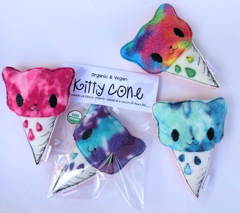 May include: Three handmade catnip toys shaped like ice cream cones. Each cone is a different color: pink, blue, and rainbow. The cones are made of fleece and have a cat face on the top. The text on the packaging reads "Organic & Vegan Kitty Cone Handmade of fleece, organic catnip & a touch of fiber fill. USDA Organic."