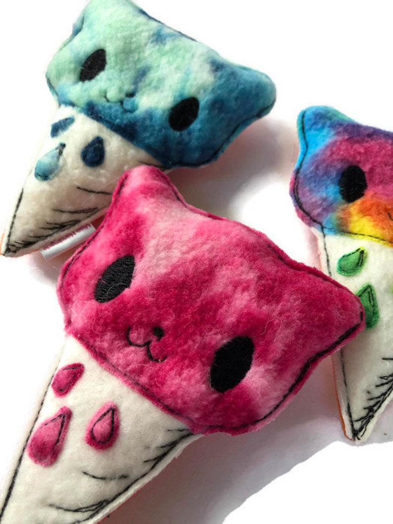 Puede incluir: Tres juguetes de peluche en forma de cono de helado con caras de animales. Los conos son blancos con costuras negras. El helado es rosa, azul y tie-dye arco&iacute;ris.