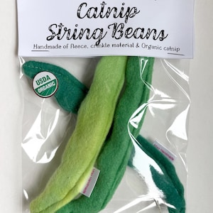 May include: A clear plastic bag containing cat toys shaped like green string beans. The bag has a white label with the text "Catnip String Beans" and "USDA Organic" seal. The toys are handmade of fleece and crinkle material.