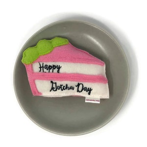 May include: A pink and white felt cake slice with green frosting and the text "Happy Dotcha Day" embroidered on it. The cake slice is on a grey plate.