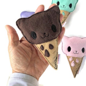 Puede incluir: Un cono de helado de fieltro marr&oacute;n, rosa y verde con una cara de gato en cada bola. Los conos de helado son hechos a mano y tienen una etiqueta que dice "Miko Handmade".