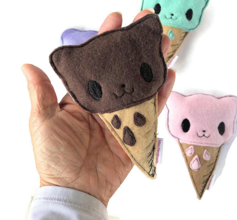 Puede incluir: Un cono de helado de fieltro marr&oacute;n, rosa y verde con una cara de gato en cada bola. Los conos de helado son hechos a mano y tienen una etiqueta que dice "Miko Handmade".