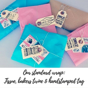 Puede incluir: Cuatro regalos envueltos en papel de seda, atados con cuerda y decorados con etiquetas estampadas a mano. Las etiquetas tienen mensajes positivos como "You are Awesome" y "Smile! Today is a new & beautiful day".