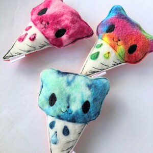 May include: Three plush ice cream cone toys with tie-dye patterns in pink, rainbow, and blue. Each cone has a cute cartoon cat face with black eyes and a white mouth.